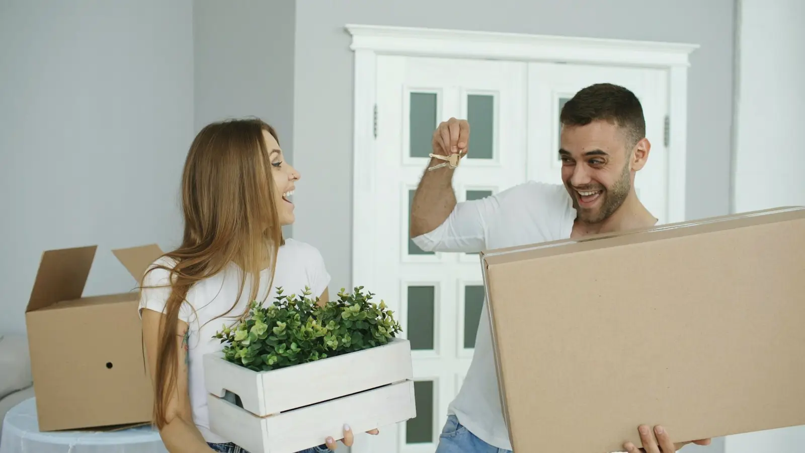 A smiling couple carrying moving boxes and plants into their new home.