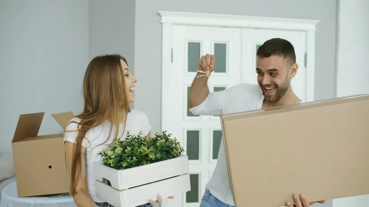A smiling couple carrying moving boxes and a crate of plants into their new home.