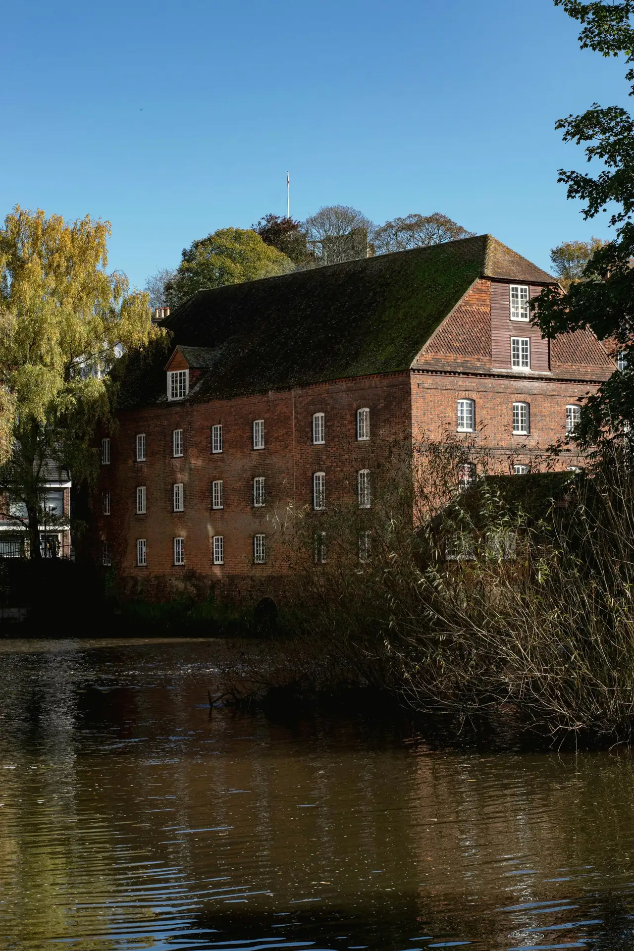 A large old red-brick mill building reflected in a calm river, trees along the bank.